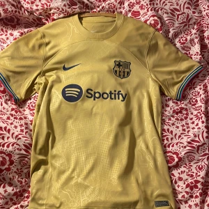 Guld fotbollströja från Nike - Säljer en snygg guld fotbollströja från Nike med FC Barcelonas emblem och Spotify-logga på bröstet. Tröjan har korta ärmar med färgglada detaljer vid ärmsluten. Perfekt för alla Barca-fans! 💛🔵🔴