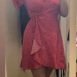 Röd prickig omlottklänning - Röd omlottklänning med vita prickar och knytband i midjan. Klänningen har korta ärmar och en feminin v-ringning.