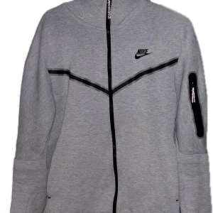Nike tech  - Snygg grå hoodie från Nike med dragkedja och svarta detaljer. Perfekt för en sportig look. Den har en praktisk ficka på ärmen och Nike-loggan på bröstet.