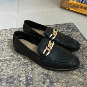 Svarta loafers med gulddetaljer - Stilrena svarta loafers från H&M med guld detalj. Använd en gång. Storlek 37. Några små trådar på insidan (se sista bilden) annars inga defekter alls och ser oanvända ut