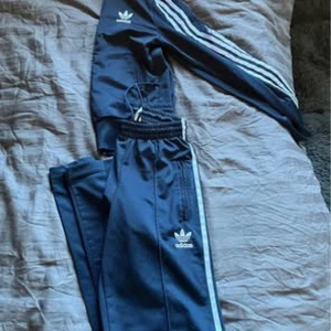 Svart träningsset från Adidas - Säljer ett snyggt svart träningsset från Adidas med de klassiska tre ränderna längs ärmar och ben. Setet består av en långärmad tröja och matchande byxor, perfekt för träning eller en avslappnad dag. Adidas-loggan syns tydligt på både tröjan och byxorna.