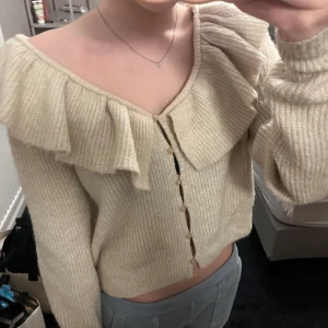Beige stickad kofta med volang - Säljer en söt beige stickad kofta med volangdetaljer och knappar framtill. Koftan har en offshoulder-design som ger en trendig look. Den är från H&M och är super snygg! 