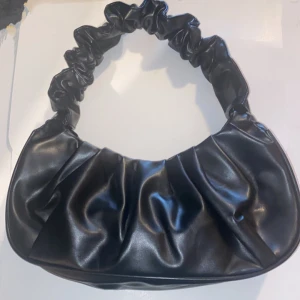 Svart axelväska i syntet - Snygg svart axelväska i syntetmaterial med rynkad design. Perfekt för att ge din outfit en trendig touch. Väskan har en unik axelrem som ger extra stilpoäng.