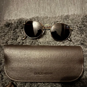 Runda solglasögon från Giorgio Armani - Snygga runda solglasögon från Giorgio Armani med mörka linser och en tunn, elegant båge. Bågarna har en subtil mönstring som ger en exklusiv känsla. Perfekta för en stilren look.