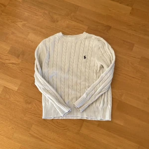 Vit stickad tröja från Ralph Lauren - Säljer en klassisk vit stickad tröja från Ralph Lauren med kabelstickat mönster och den ikoniska loggan broderad på bröstet. Tröjan har långa ärmar och en rund halsringning. Perfekt för en stilren look. Den är rätt så löddrig.