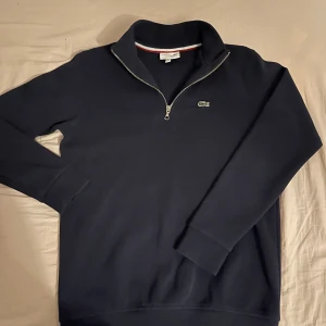 Mörkblå tröja med dragkedja från Lacoste - Säljer en stilren mörkblå tröja från Lacoste med halv dragkedja och broderad logotyp på bröstet. Tröjan har långa ärmar och en ribbad krage med röd och vit detalj på insidan. Perfekt för en avslappnad och snygg look.