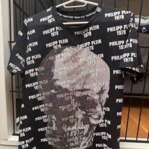 Svart t-shirt med dödskalle från Philipp Plein - Cool svart t-shirt från Philipp Plein med ett stort dödskalletryck i silver på framsidan. T-shirten har ett all-over print med texten 'Philipp Plein 1978'. Perfekt för en edgy look!