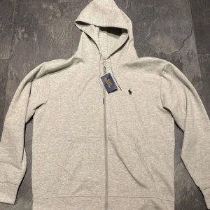 Grå hoodie från Ralph Lauren - Säljer en stilren grå hoodie från Ralph Lauren med dragkedja och den klassiska loggan broderad på bröstet. Tröjan har en bekväm passform och är perfekt för kyligare dagar. Den har långa ärmar och en praktisk huva.