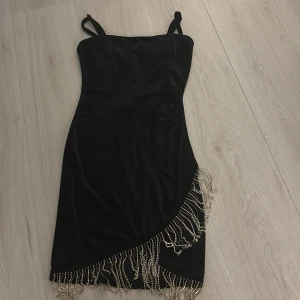 Svart klänning med pärldetaljer - Elegant svart klänning med smala axelband och pärldetaljer längs fållen. Perfekt för en festlig kväll med sin stilrena design och unika detaljer.