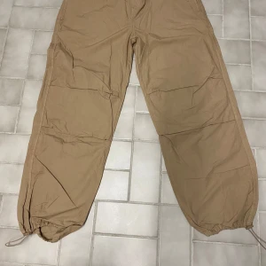 Beige cargopants med snörning - Säljer ett par beige cargopants med avslappnad passform. Byxorna har praktiska snörningar vid bensluten och flera fickor för en cool och funktionell stil. Perfekta för en casual look.