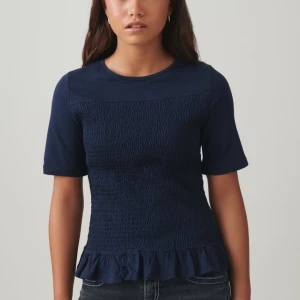 Mörkblå topp med volangdetalj - Snygg mörkblå topp med korta ärmar och volangdetalj vid nederkanten. Toppen har en smockad textur på framsidan som ger en unik look. Perfekt för att piffa upp vardagsstilen. storlek 158/164 men skulle säga att den sitter som xs-s som jag alltid har😇😇 använd en gång, och den är slut på hemsidan