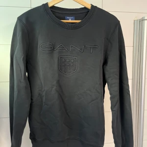 Svart tröja från GANT - Snygg svart tröja från GANT med broderad logotyp och emblem på bröstet. Tröjan har långa ärmar och ribbad halsringning. Perfekt för en stilren look. Storlek S. 