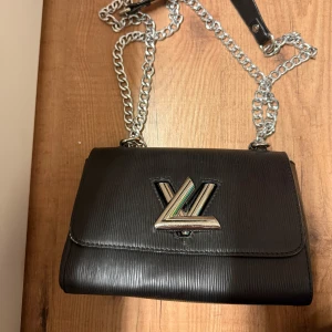 Svart axelväska med silvriga detaljer 🤍 - Elegant svart axelväska med en stilren design och en kedjerem. Väskan har en distinkt V-detalj på framsidan och är tillverkad i skinn med en texturerad yta. Perfekt för att ge en lyxig touch till din outfit.