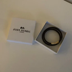 Svart armband från John Henric - Elegant svart/brunt armband från John Henric med gulddetalj framtill. Perfekt för en stilren look. Helt nytt aldrig använt.