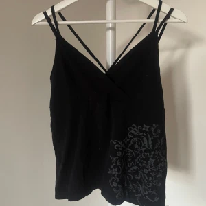 Svart omlottlinne med mönster - Snyggt svart omlottlinne med smala axelband och ett diskret mönster på sidan. Perfekt till träning eller till vardags. Väldigt skönt material. Köpt på H&M för väldigt länge sedan. Från H&M athletic.