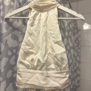 Vit halterneck topp med knytband - Snygg vit halterneck topp med knytband i midjan. Perfekt för en stilren look. Toppen har en elegant design med en hög hals och ärmlös stil.