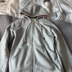 Grå hoodie från Burberry - Säljer en stilren grå hoodie från Burberry med klassiskt rutigt mönster i huvan. Tröjan har dragkedja framtill och praktiska fickor. Perfekt för en avslappnad och trendig look. Använd ett fåtal gånger men ser helt ny ut!