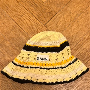Mössa/hatt  från Ganni - Snygg randig hatt från Ganni i bomull med gula, svarta och vita ränder. Perfekt för sommaren med sin lätta och luftiga design. Hatten har en bred kant och ett broderat Ganni-märke framtill.