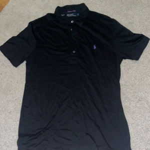 Svart pikétröja från Ralph Lauren - Snygg svart pikétröja från Ralph Lauren med klassisk krage och knappar framtill. Den har en liten lila logga broderad på bröstet. Perfekt för en stilren look.