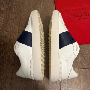 Valentino skor - Snygga vita Valentino Garavani sneakers med breda svarta band över ovansidan. Skorna har en klassisk design med en robust sula och kommer med en röd förvaringspåse.