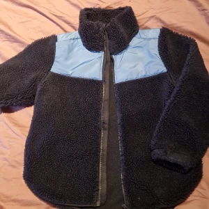 Svart teddyjacka med blå detaljer - Mysig svart teddyjacka från Arket med ljusblå detaljer på axlarna. Jackan har en dragkedja framtill och är perfekt för kyligare dagar. Den är långärmad och har en hög krage för extra värme.