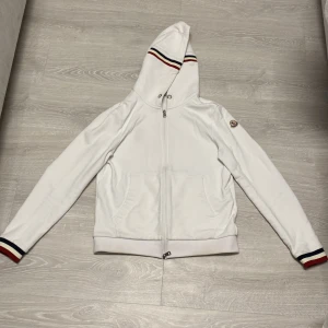 Moncler kofta - Säljer en stilren vit huvtröja från Moncler med dragkedja och broderad logga på ärmen. Tröjan har randiga detaljer i rött och blått på huvan och ärmsluten. Perfekt för en casual look!