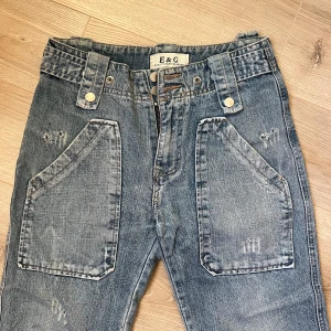 Blå jeans från E&G - Snygga blå jeans från E&G med unika fickdetaljer och slitningar. Byxorna har en rak passform med stora fram- och bakfickor med knappar. Perfekta för en avslappnad stil.