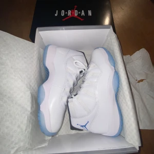 Nike Air Jordans i Legend blue - Snygga Nike Air Jordans i vitt med ljusblå detaljer och svart insida. Skorna har en hög design med snörning och den ikoniska Jumpman-loggan på hälen. Perfekta för dig som vill ha stil och komfort i ett.