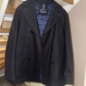 Svart kappa från Tommy Hilfiger - Snygg svart kappa från Tommy Hilfiger med dubbelknäppning och klassisk krage. Jackan har en quiltad insida för extra värme och stilrena fickor framtill. Perfekt för kyligare dagar.