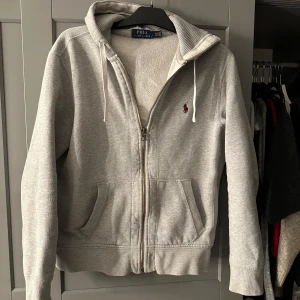 Grå Polo Ralph Lauren hoodie - Säljer en stilren grå hoodie från Polo Ralph Lauren. Tröjan har en bekväm passform med långa ärmar och en praktisk huva. Har en liten fläck på luvan men inget man tänker på.