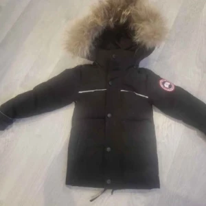 Svart dunjacka från Canada Goose - Säljer en svart dunjacka från Canada Goose med pälskantad huva. Jackan har en klassisk design med knappar och dragkedja framtill samt en logotyp på ärmen. Perfekt för kalla vinterdagar.   OBS BARN JACKA ca 98 i vanlig strl!