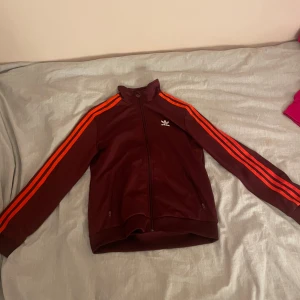 Vinröd träningsjacka från Adidas - Snygg vinröd träningsjacka från Adidas med klassiska tre ränder i orange längs ärmarna. Jackan har dragkedja framtill och en stor vit Adidas-logga på ryggen. den är i storlek s eller xs lite oklart men hör av er provat om ni vill ha bild på! är osäker på om det är design för tjejer eller killar men skulle tro tjejer. använd ganska många gånger.