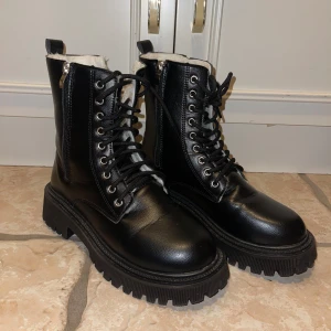 Svarta kängor med snörning och dragkedja - Snygga svarta kängor i skinn med både snörning och dragkedja för enkel på- och avtagning. De har en robust sula och är fodrade med vitt material för extra värme. Perfekta för kyligare dagar.