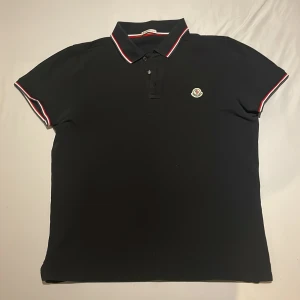 Svart pikétröja från Moncler - Snygg svart pikétröja från Moncler med klassisk krage och knappar framtill. Tröjan har korta ärmar med röd och vit randdetalj vid ärmslut och krage. Perfekt för en stilren look.