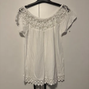 Vit blus med spetsdetaljer från ONLY - Säljer en söt vit blus från ONLY med vackra spetsdetaljer vid halsringningen och nederkanten. Blusen har korta ärmar och en luftig passform, perfekt för varma dagar. Passar både till jeans och kjol för en avslappnad look.