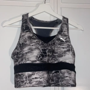 Svart och vit sport-bh från Puma - Snygg sport-bh från Puma i svart, vitt och grått med ett abstrakt mönster. Den har en racerback-design och bred resår under bysten för extra stöd. Använd enstaka gånger.
