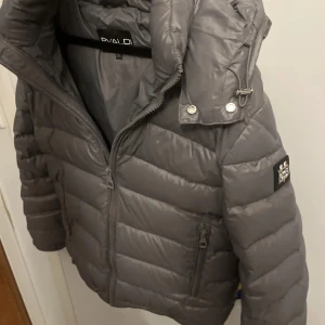 Grå pufferjacka från BValdi - Snygg grå pufferjacka från B. Valdi med dragkedja och hög krage. Jackan har en quiltad design och praktiska fickor med dragkedjor. Riktigt bekväm jacka! Pris går att diskutera storlek M men passar S extremt bra också 