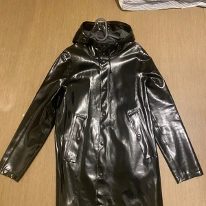 Stutterheim regnjacka - Snygg svart regnjacka från Stutterheim med huva och knappar framtill. Jackan är glansig som gör även att jackan är väldigt unik. Storleken är xxs men passa som xs eftersom de är stora i storlek.