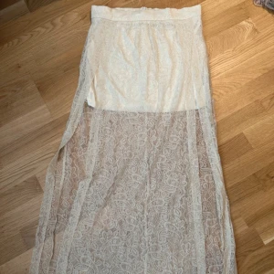 Vit spetskjol från Bik Bok - Säljer en vit långkjol från Bik Bok i storlek M. Kjolen har ett vackert spetsöverdrag och en underkjol i satinliknande material. Perfekt för en stilfull look.