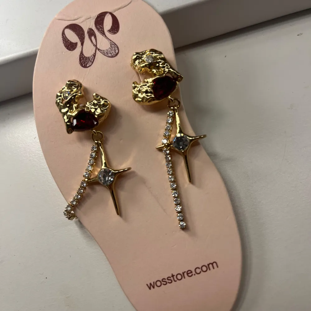 Eleganta guldörhängen från WOS med röda hjärtan och gnistrande stjärndetaljer. De har en hängande design med strass som ger en extra touch av glamour. Perfekta för att lysa upp vilken outfit som helst.. Asusteet.