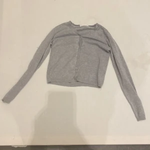 Grå kofta från KappAhl - Säljer en stilren grå kofta från KappAhl. Den har långa ärmar och knappar framtill, perfekt för lager-på-lager-stil. Enkel och klassisk design som passar till många tillfällen.