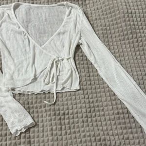 Vit omlottopp med knytband - Säljer en stilren vit omlottopp med långa ärmar och knytband i midjan. Toppen är ribbad och croppad, perfekt för en trendig look. Om ni vill se den på, skriv gärna!