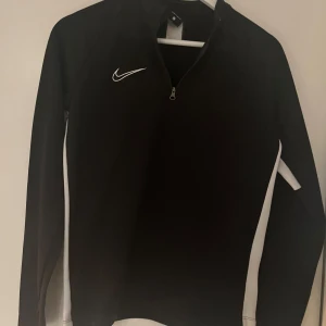 Svart långärmad tröja från Nike - Säljer en svart långärmad tröja från Nike med vit logga och dragkedja vid halsen. Tröjan har vita detaljer längs sidorna och är tillverkad i Dri-FIT-material för optimal komfort.