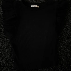 Svart ribbad topp från Zara - Snygg svart ribbad topp från Zara med volangdetaljer på axlarna. Säljs inte längre 