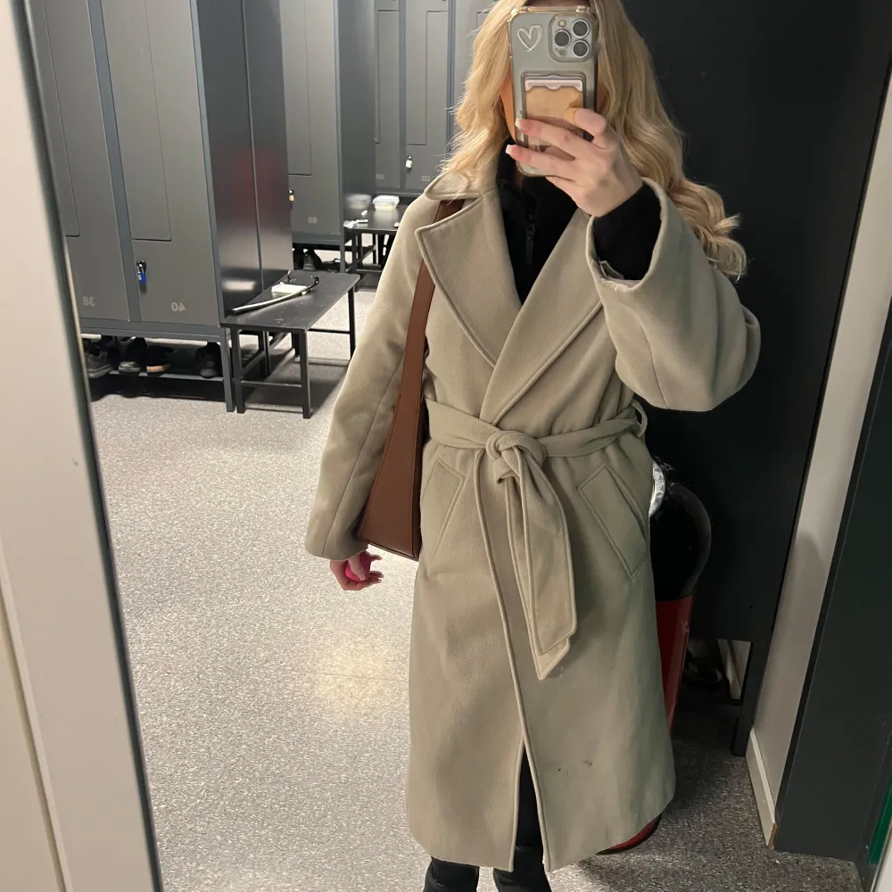 Snygg beige kappa med knytband i midjan. Perfekt för kyligare dagar med sin klassiska design och långa ärmar. Tung kappa så känns dyr när man har på sig den. Har små fläckar på framsidan därav priset. Köptes för 899 kr. Kappan säljs inte längre.. Takit.