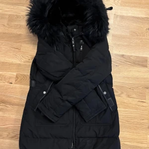 Svart dunjacka från Cedrico - Snygg svart dunjacka från Cedrico med pälskantad huva och dragkedja. Jackan har praktiska fickor med knappar och justerbara remmar i midjan för en perfekt passform. Använd 1 vinter. Jackan är hel och ren. Äkta päls på hela jackan.  OBS PÅ 4de bilden syns dragkedjans fäste längst ner, en liten söm av den gick upp så har sytt fast den. Det är ingenting som syns när man har på sig jackan utan synd 