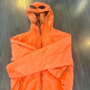 Cp Company jacka - Säljer en cool orange dunjacka från C.P. Company. Jackan har en unik design med inbyggda glasögon i huvan och dragkedja framtill. Perfekt för att sticka ut i mängden och hålla sig varm under kalla dagar.