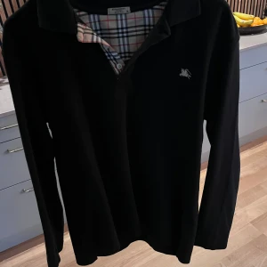 Svart pikétröja från Burberry - Säljer en  svart pikétröja från Burberry med klassiskt rutigt mönster på insidan av kragen. Tröjan har en broderad logotyp på bröstet. 