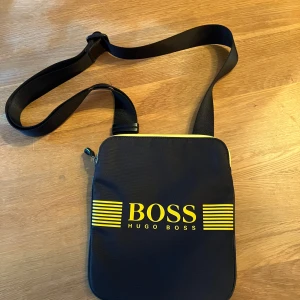 Svart axelväska från Hugo Boss - Snygg svart axelväska från Hugo Boss med gul logotyp och detaljer. Väskan har en justerbar axelrem och dragkedja upptill. 
