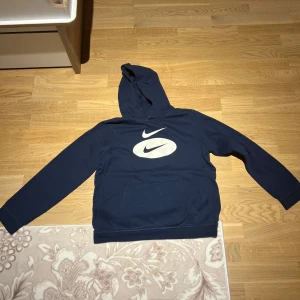 Mörkblå hoodie från Nike - Säljer en mörkblå hoodie från Nike med det klassiska logotyptrycket i vitt på framsidan. Tröjan har en bekväm passform med långa ärmar och en praktisk känguruficka. Perfekt för en avslappnad stil.